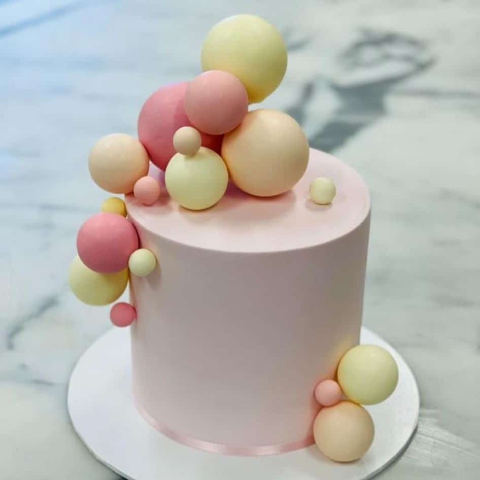 Custom decorated cake available at Brunetti Classico Carlton. Pink, orange and yellow balloon or chocolate sphere decoration on a light pink cake