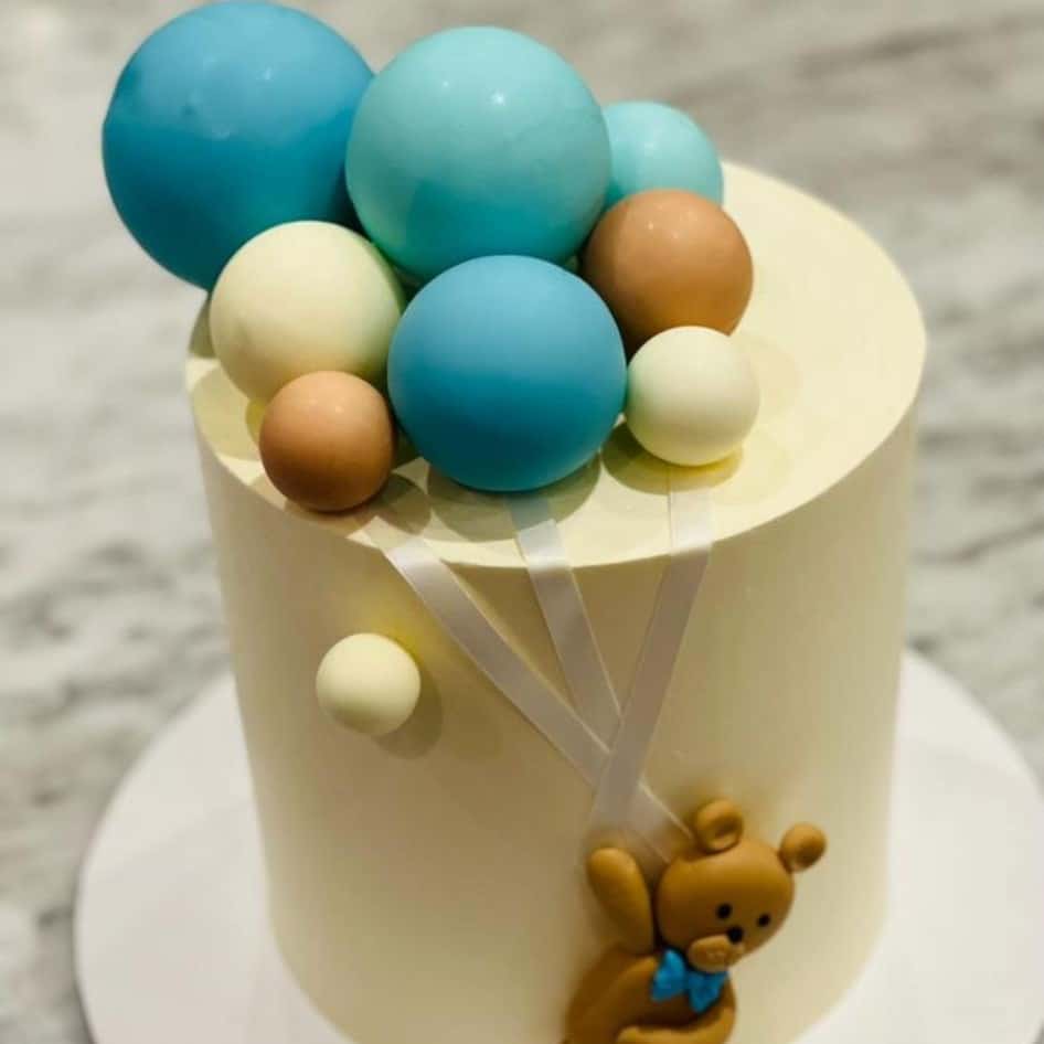 Custom decorated cake available at Brunetti Classico Carlton. Boys 1st birthday party cake with teddy bear and balloon decorations. Chocolate sphere decoration with blue, white and light brown balloons. on a light yellow cake. Baby shower cake, gender reveal cake, teddy bear cake