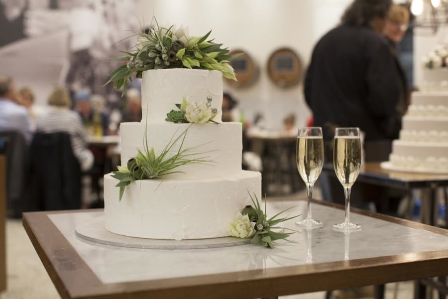 Three-tier white wedding cake with greenery and flowers beside champagne glasses.