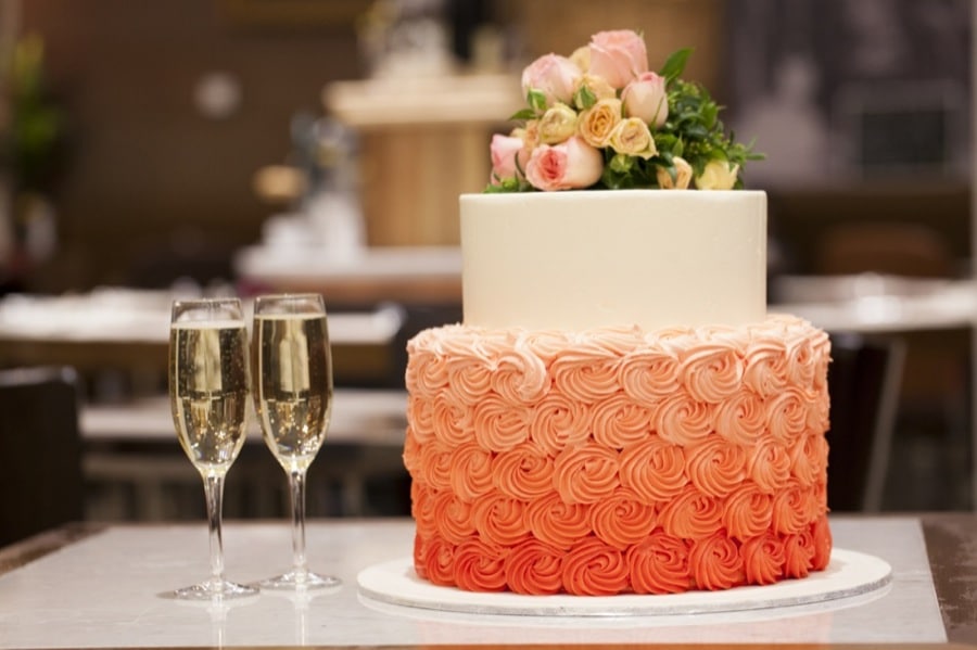 Two-tier cake with ombre orange frosting and floral top, next to two champagne glasses.