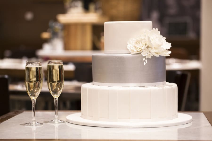 Three-tier white and silver wedding cake with floral decoration beside two champagne glasses.